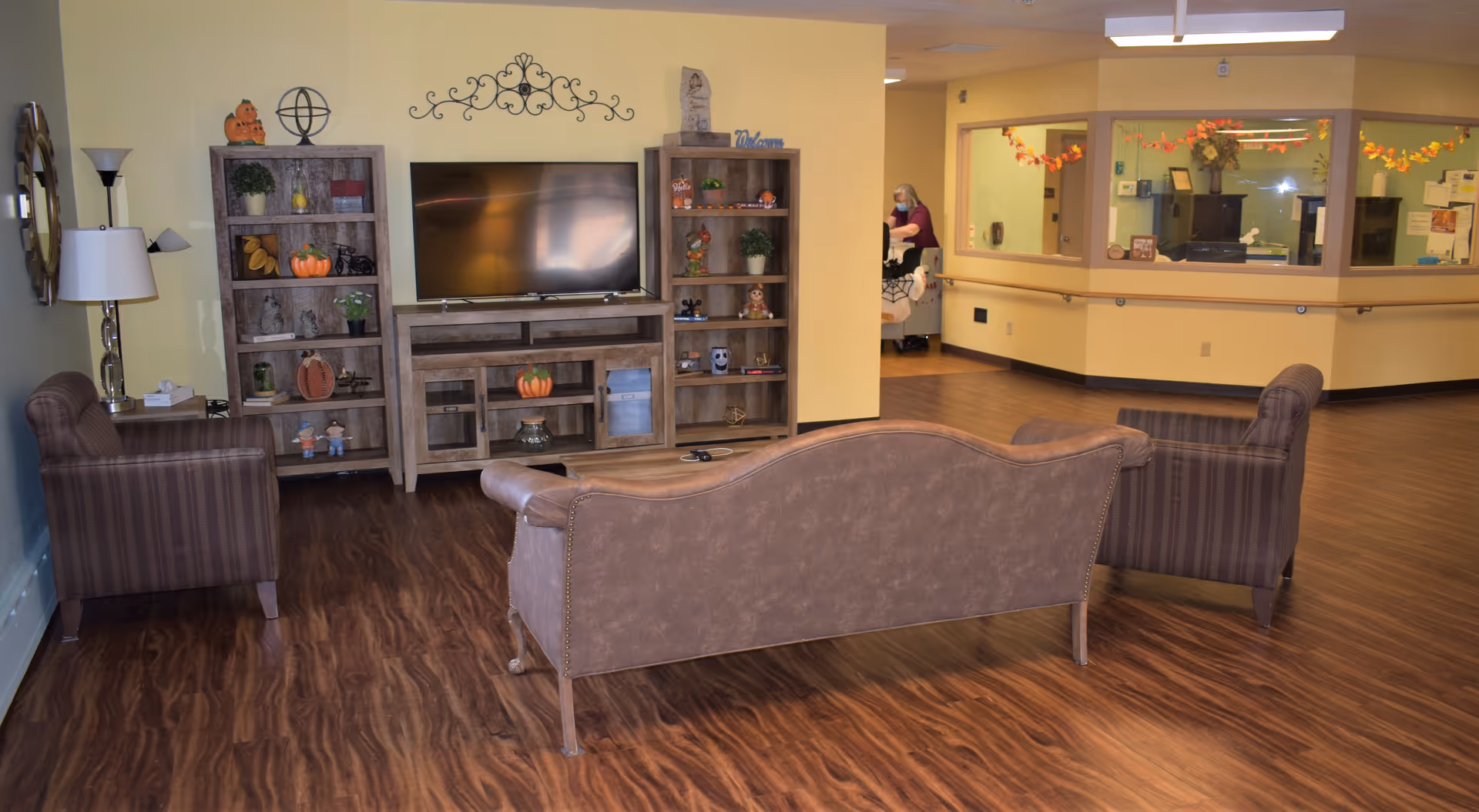 A cozy living room area in a senior care facility with a brown leather sofa and two striped armchairs arranged around a wooden TV stand and two matching bookshelves. The shelves are decorated with small plants, pumpkins, and other seasonal decor. The floor is wood laminate, and there is a reception area visible in the background with a staff member wearing a mask.