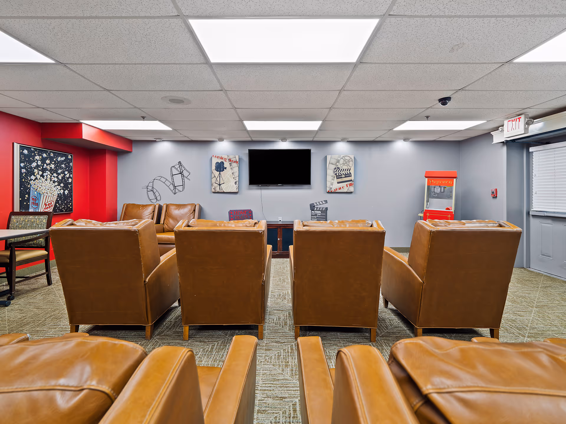 A senior living media room with rows of brown leather chairs facing a wall-mounted TV, movie-themed decor, and a popcorn machine.