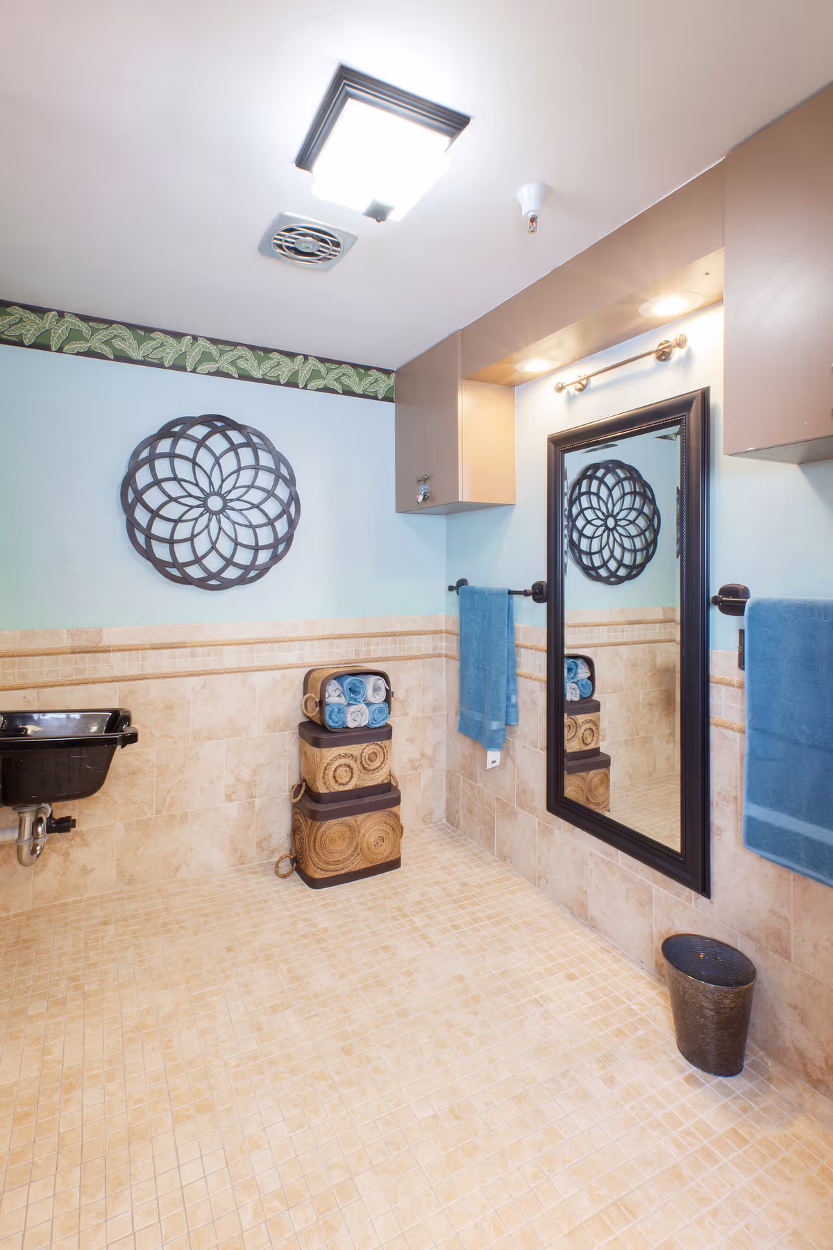 A clean bathroom with beige tiled floor and walls, a black wall-mounted sink, a large rectangular mirror with a black frame, two blue towels hanging on towel racks, decorative wall art, and stacked wicker baskets holding rolled towels.