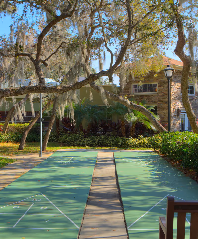 Outdoor shuffleboard courts with green playing surfaces and white markings, surrounded by trees with hanging moss and lush landscaping. A stone building with windows and a lamp post is visible in the background under a clear blue sky.