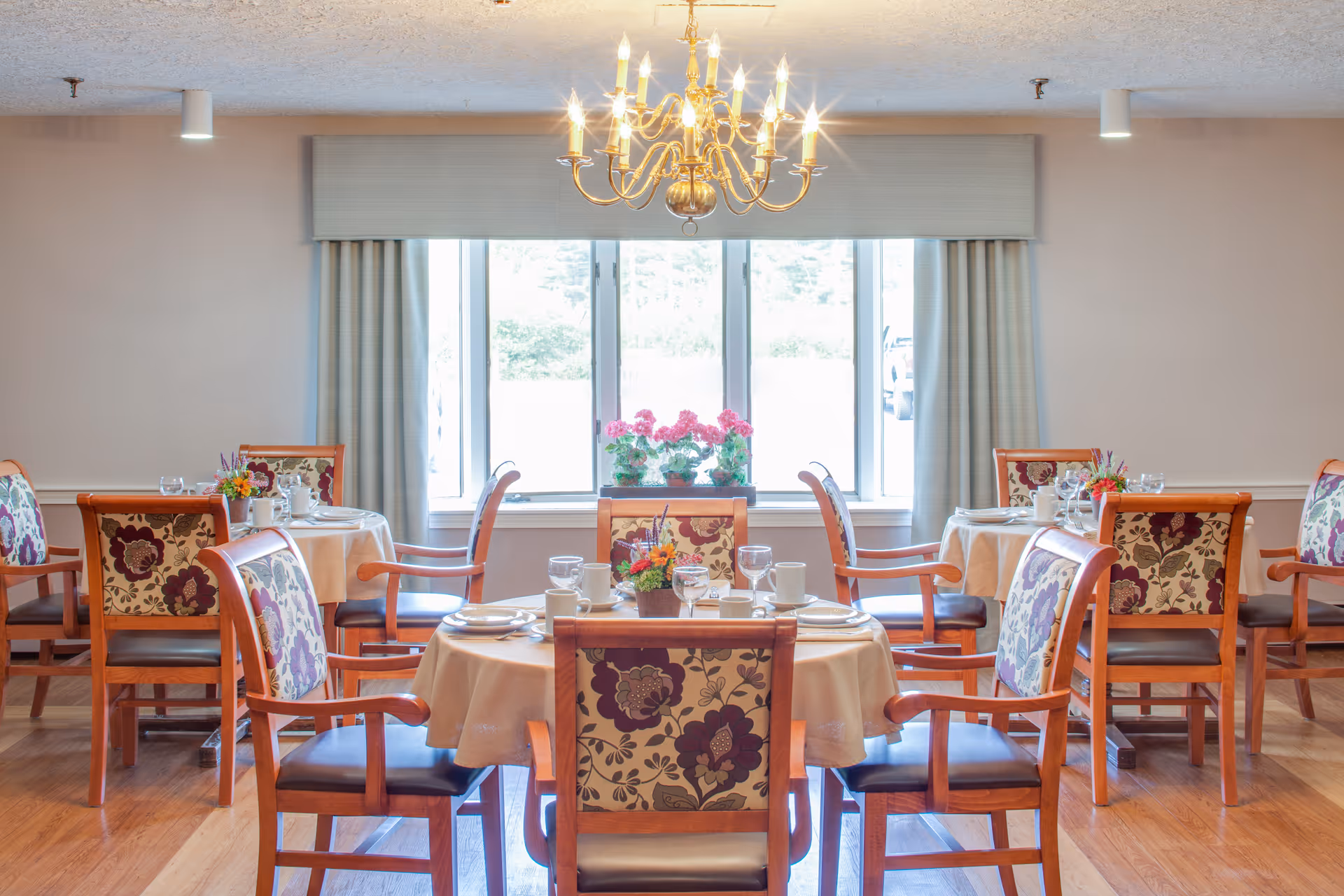 Bright dining room with round tables set for meals and floral-upholstered wooden chairs beneath a chandelier and large window.
