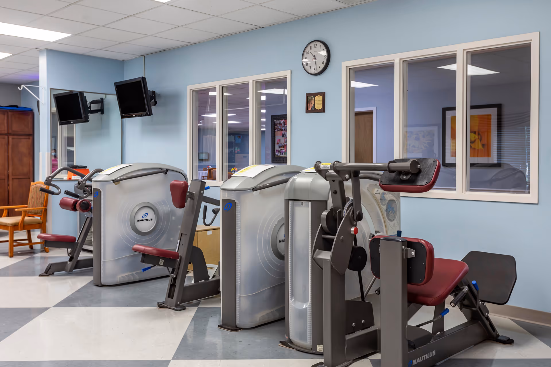 Interior view of a fitness room in a rehabilitation and health care center featuring several Nautilus exercise machines with maroon seats and handles, light blue walls, a clock, and two wall-mounted televisions.