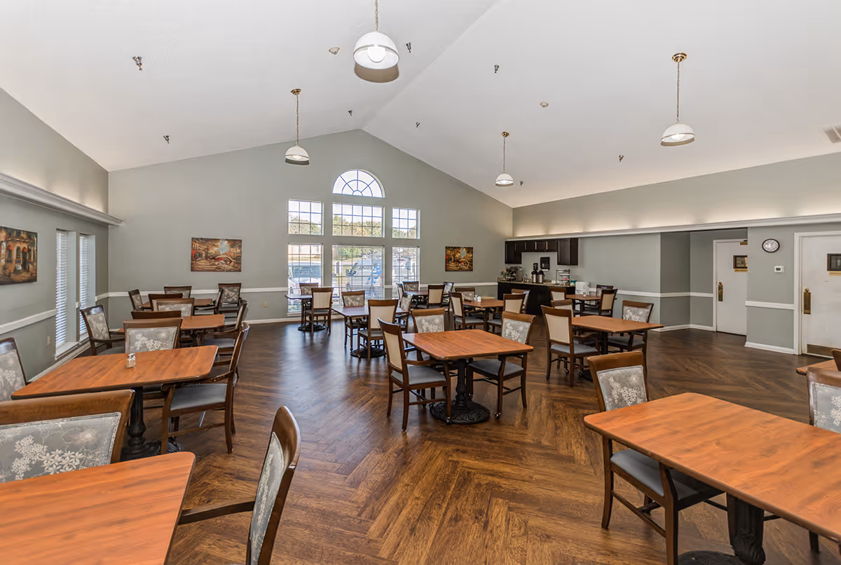 A spacious dining room with multiple wooden tables and chairs arranged neatly. The room has a high vaulted ceiling with hanging pendant lights, large windows letting in natural light, and paintings on the walls. There is a counter area with coffee machines and supplies in the back corner.