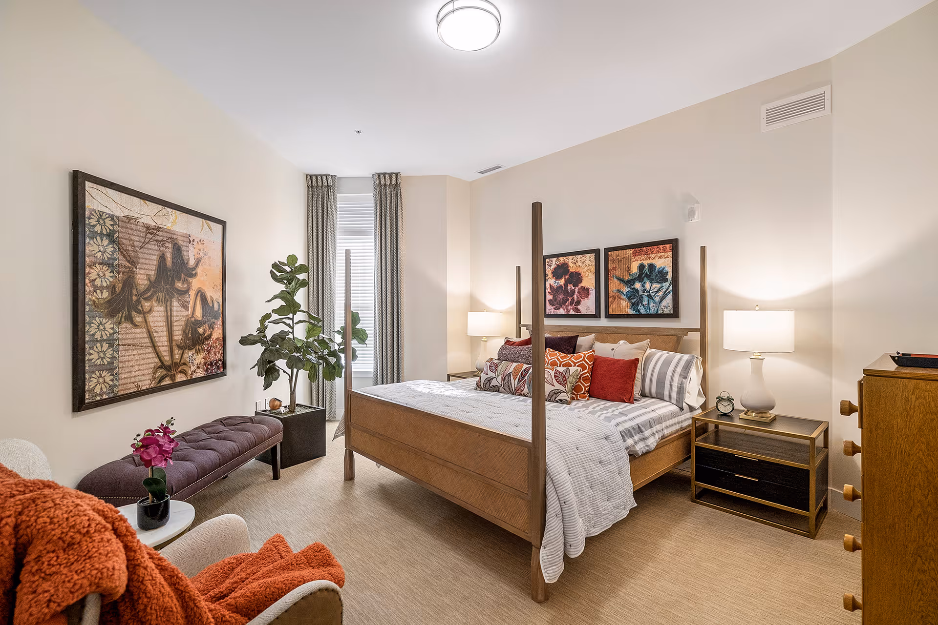 A cozy bedroom with a wooden four-poster bed adorned with multiple decorative pillows and a striped bedspread. On either side of the bed are nightstands with lamps and an alarm clock on the right nightstand. The room also features a wooden dresser, a purple cushioned bench, a potted plant near the window with curtains, a white armchair with an orange throw blanket, and framed floral artwork on the walls.