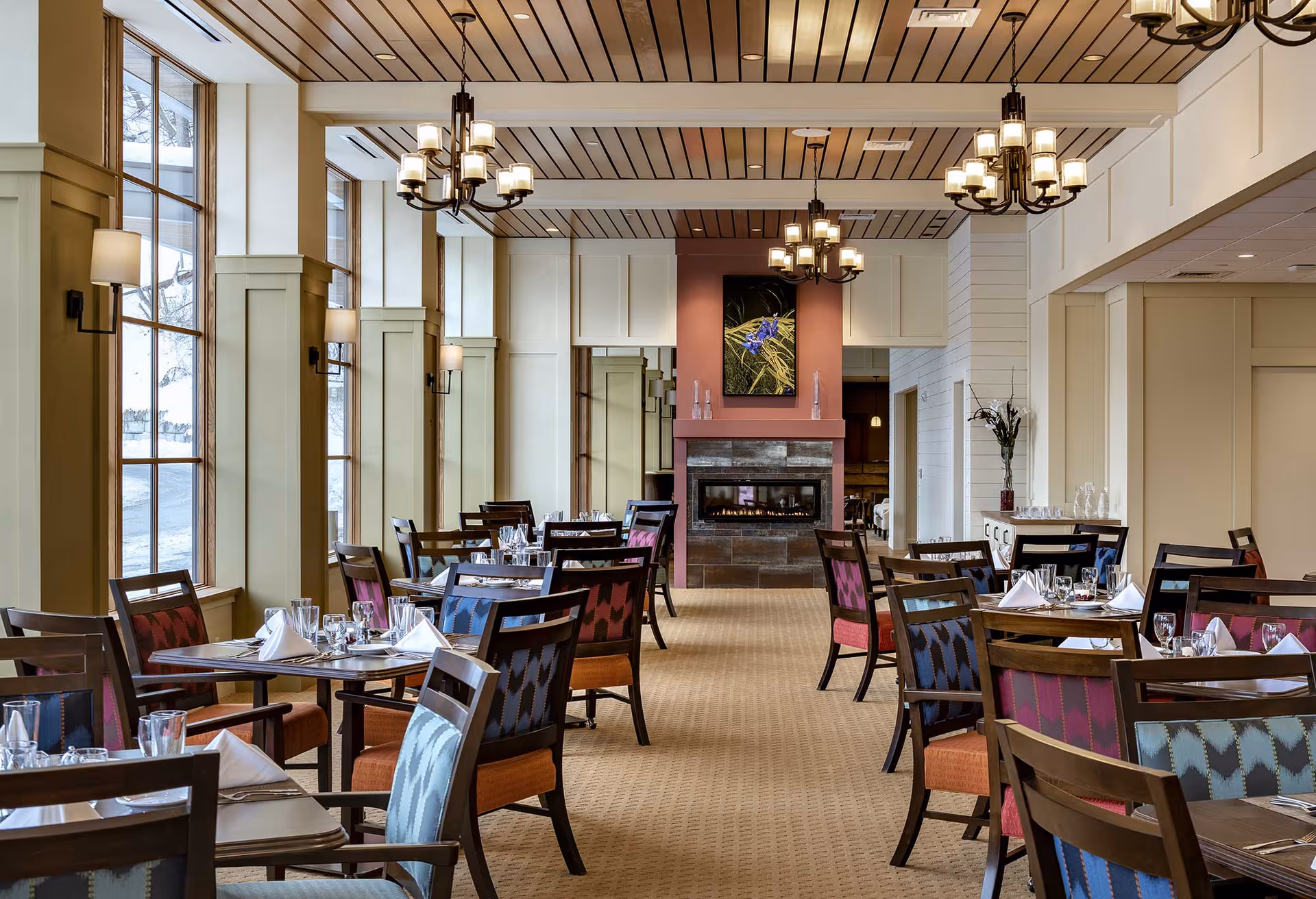 A spacious dining room with multiple tables set with white napkins, glasses, and silverware. The room features large windows on the left side letting in natural light, beige walls with paneling, and a ceiling with wooden slats and hanging chandeliers. At the far end, there is a fireplace with a framed floral artwork above it. Chairs around the tables have colorful patterned upholstery in shades of blue, red, and orange.