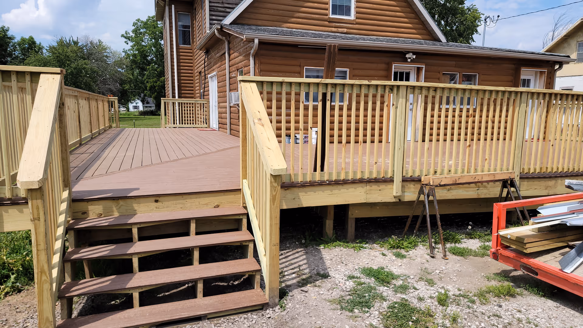 A wooden deck with stairs and railings attached to a log cabin style building. The deck is elevated with wooden steps leading down to a gravel and grass area. There is some construction material and equipment visible on the right side near the deck.