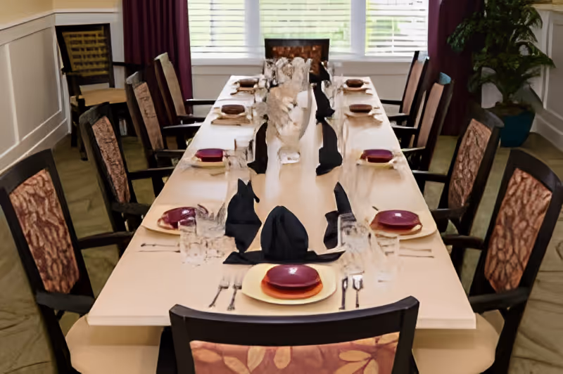 A long dining table set for a meal with multiple place settings including plates, glasses, silverware, and black folded napkins. The table is surrounded by cushioned chairs with patterned upholstery. A decorative centerpiece is placed in the middle of the table. The room has large windows with white blinds and purple curtains, and a potted plant is visible in the corner.