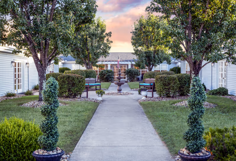 Paved walkway leading to a courtyard fountain flanked by benches, trees, and single-story buildings at sunset.