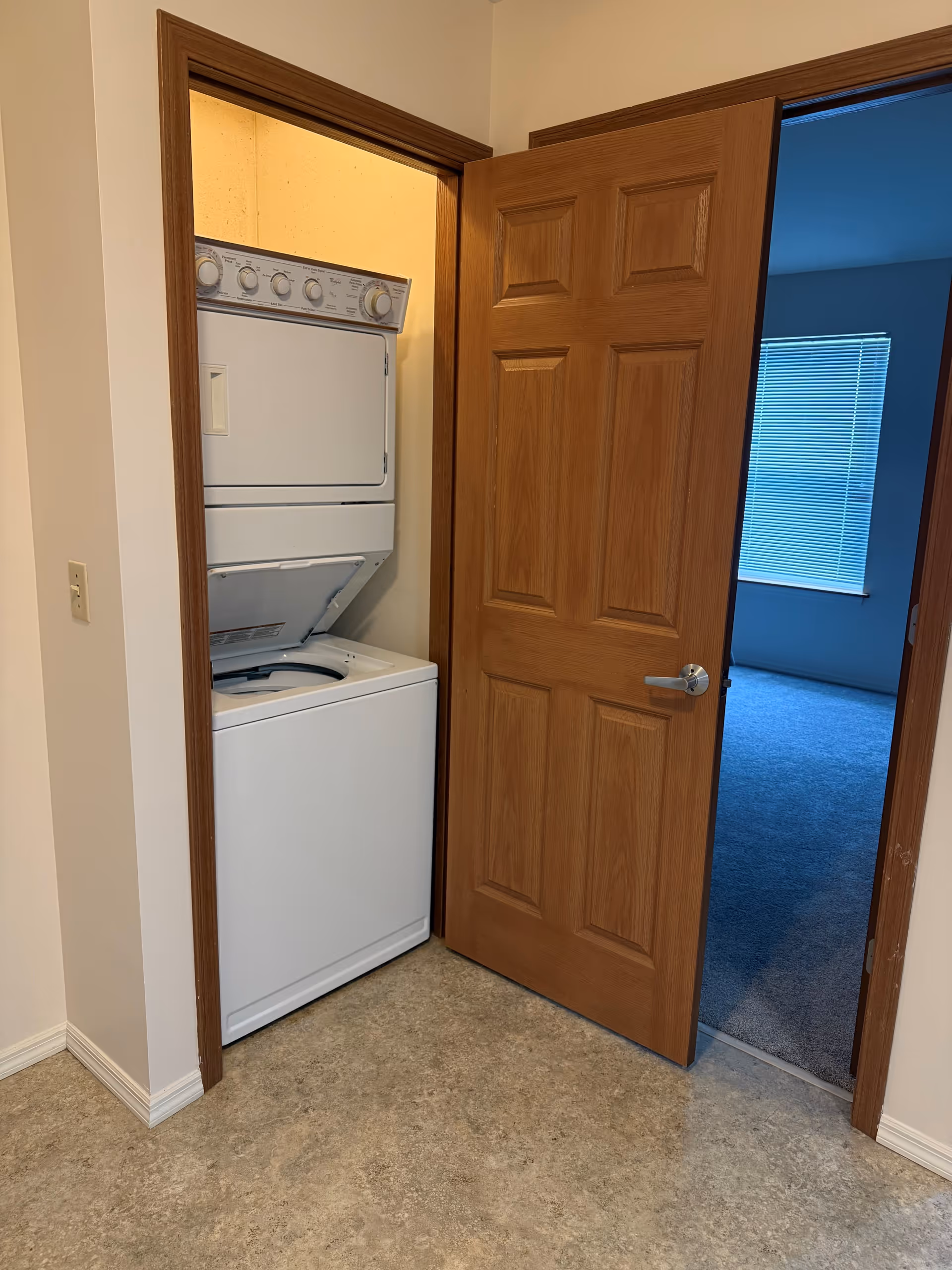 A small laundry closet with a stacked washer and dryer unit. The closet has a wooden door that is partially open, revealing a carpeted room with a window and blinds in the background.