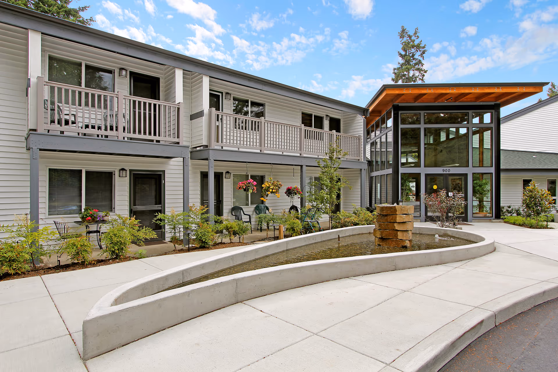 Front entrance and courtyard of Evergreen Court showing a two-story building with balconies, landscaped planters, and a curved water feature.