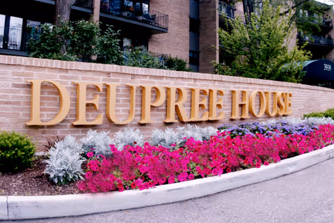 A large brick sign with gold letters spelling 'DEUPREE HOUSE' surrounded by colorful flowers and greenery, with a multi-story building and trees in the background.