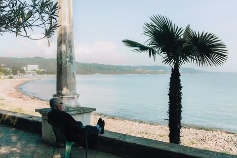 An elderly man sitting on a chair by a stone railing, looking out over a calm sea with a rocky shoreline and distant hills under a partly cloudy sky. A palm tree and other greenery are nearby.