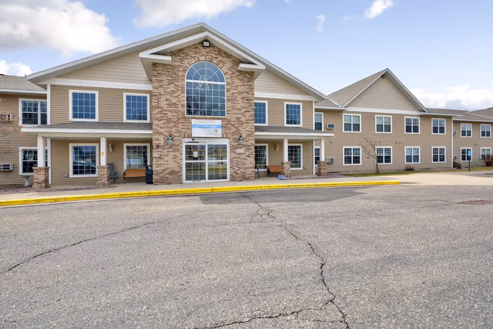 Exterior view of a two-story senior living facility building with beige siding and brick accents around the entrance. The entrance features large glass doors and a tall arched window above. There are benches on the covered porch area near the entrance, and the building is surrounded by a paved driveway and parking area under a partly cloudy sky.