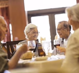 A group of elderly people sitting around a dining table, enjoying conversation and drinks in a well-lit room with large windows in the background.