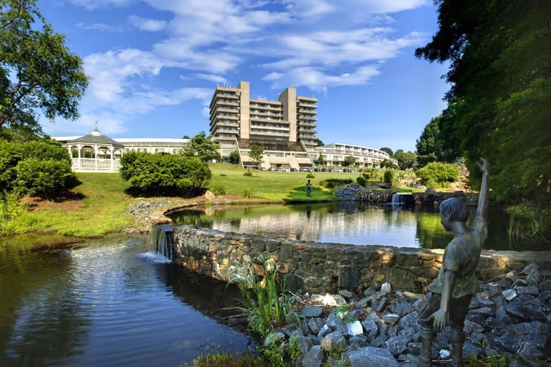 A scenic outdoor view of Cokesbury Village featuring a large multi-story building in the background, a well-maintained lawn, a pond with a small waterfall, a white gazebo, and a statue of a child waving near the water's edge, surrounded by trees and blue sky with scattered clouds.