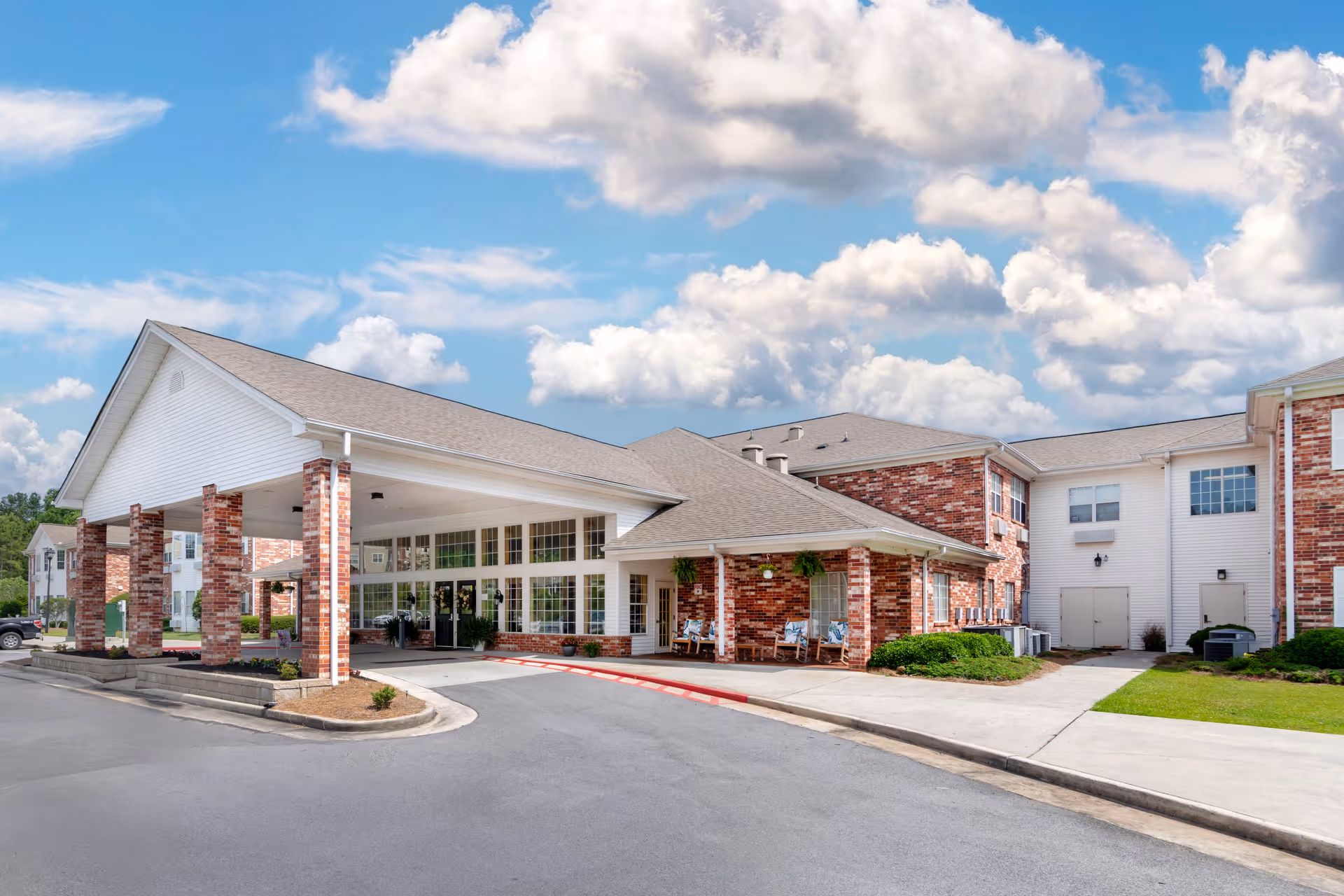 Exterior view of Brookdale Rome senior living facility showing a large covered entrance with brick pillars, multiple windows, and a driveway under a partly cloudy blue sky.