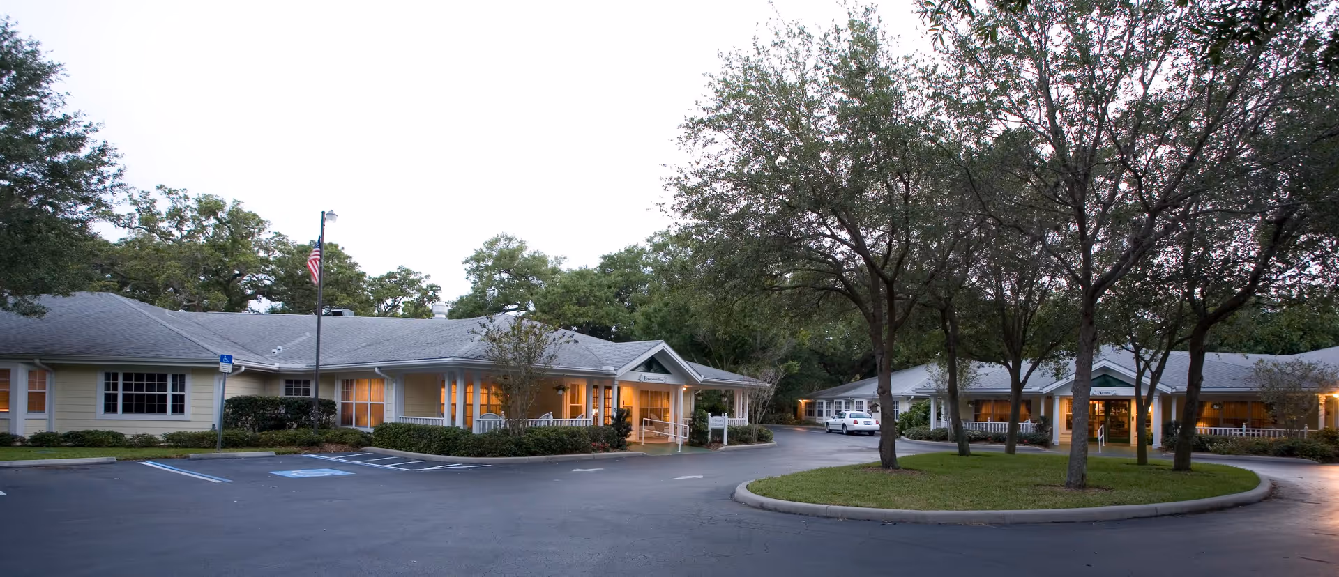 Front exterior of a single-story senior living facility with a circular driveway, parking spaces, and trees at dusk.