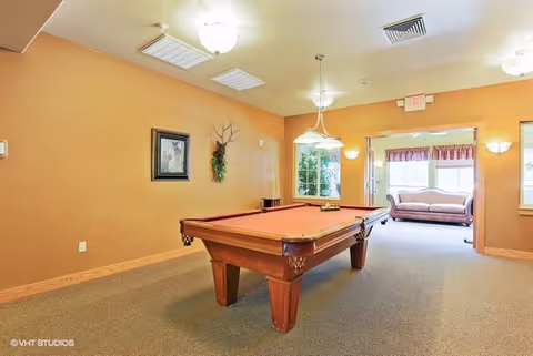 A recreational room with a pool table in the center, beige walls, and carpeted floor. There is a framed picture and a decorative wall hanging on the left wall. In the background, there is a seating area with a couch near large windows letting in natural light. Ceiling lights and a hanging light fixture illuminate the room.