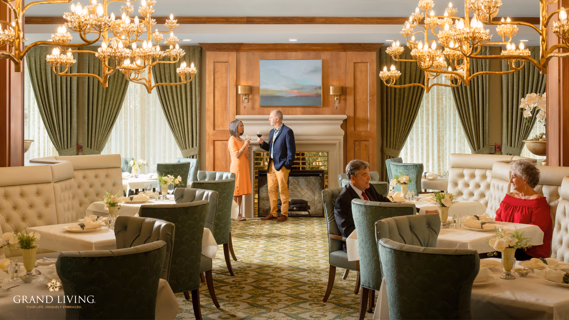 A warmly lit dining room with elegant chandeliers, green upholstered chairs, and white tufted booth seating. Two people stand near a fireplace in the background, holding wine glasses and conversing. Two other people are seated at tables, engaged in conversation. The room features large windows with green curtains and a patterned carpet.