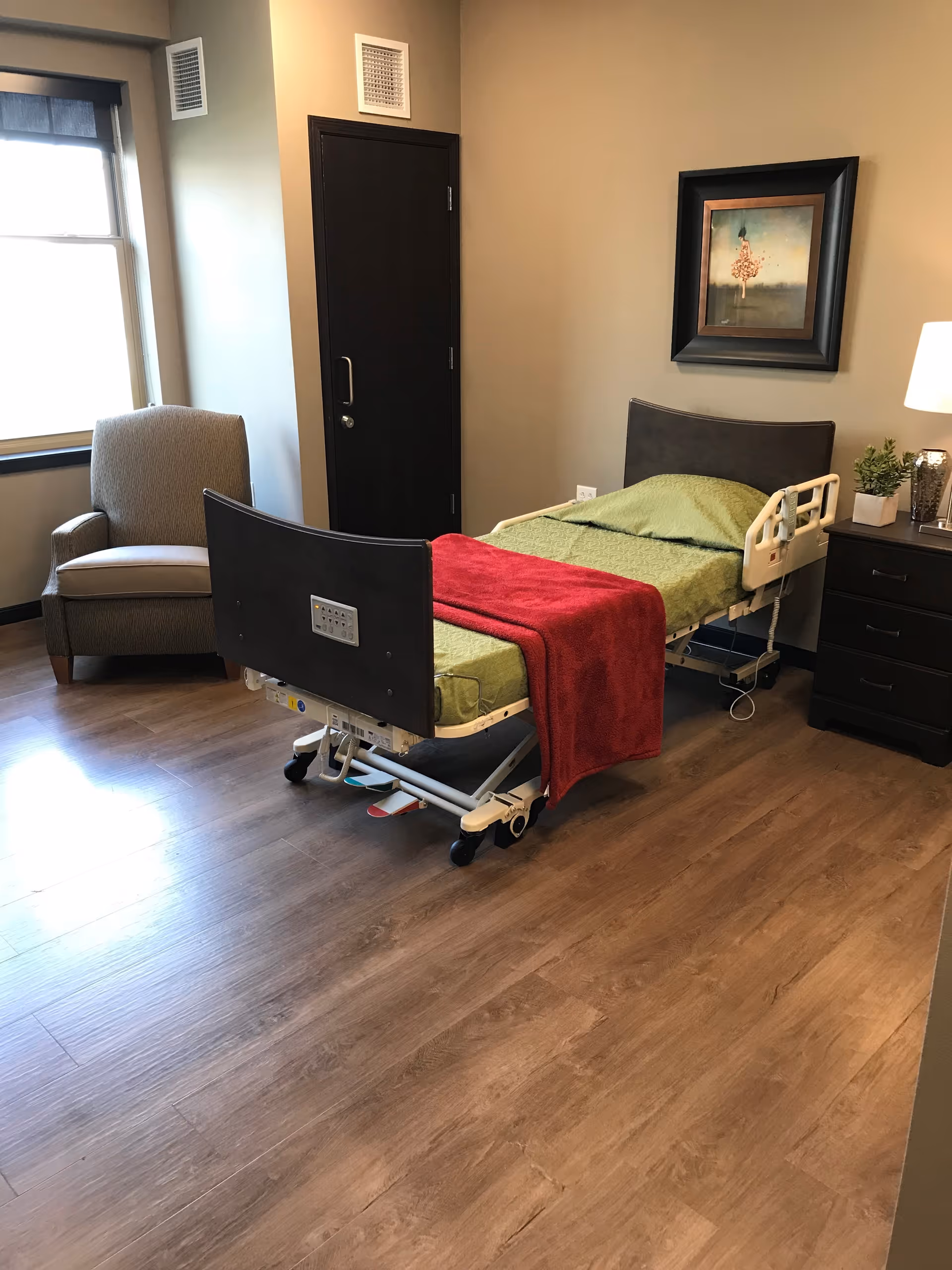 A room with a hospital-style bed covered with green bedding and a red blanket, a gray armchair near a window, a black door, a nightstand with a lamp and a small plant, and a framed picture on the beige wall.