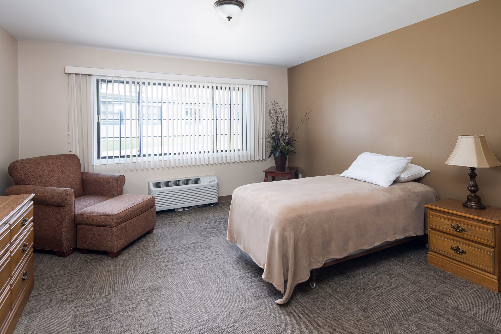 A simple bedroom with a single bed covered in a beige blanket and white pillows, a wooden nightstand with a lamp, a wooden dresser, a brown upholstered armchair with an ottoman, a large window with vertical blinds, and a small table with a plant in the corner.