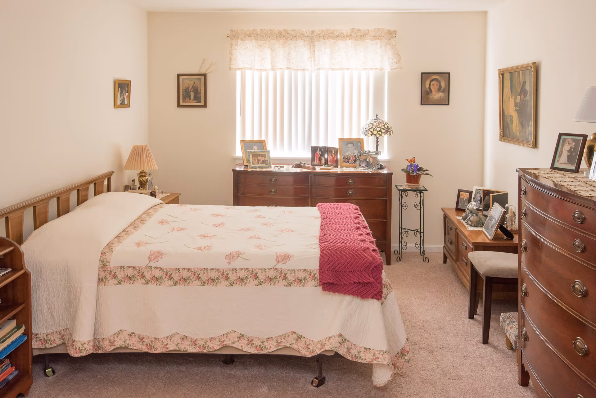 Cozy bedroom with a floral quilted bed, wooden dressers and nightstands topped with framed photos and lamps beneath a window with vertical blinds.
