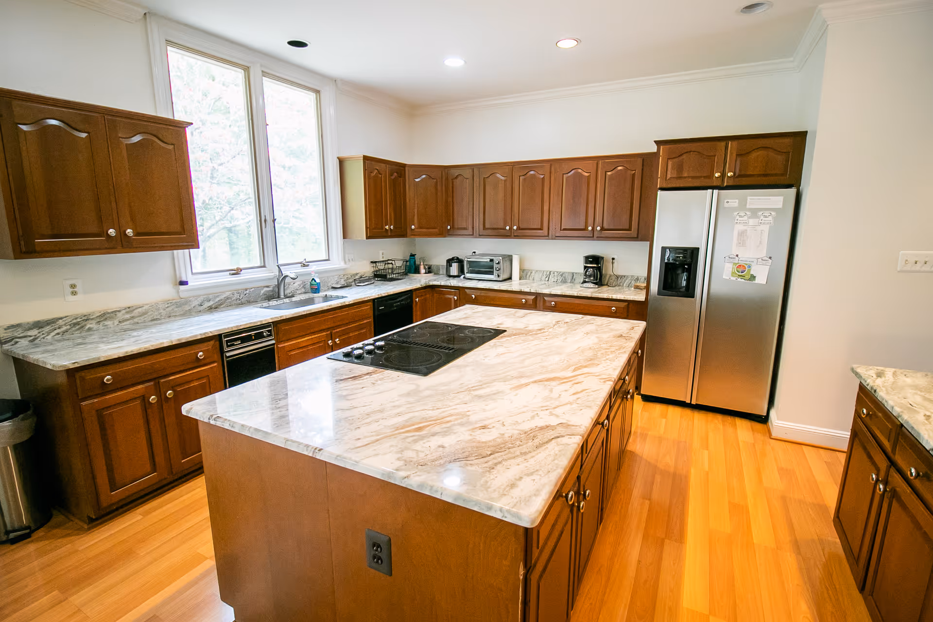 A spacious kitchen with wooden cabinets and marble countertops. The kitchen features a large central island with a built-in electric cooktop, a double-door stainless steel refrigerator, a dishwasher, a toaster oven, and a coffee maker. There are two large windows above the sink letting in natural light, and the floor is made of light-colored wood.