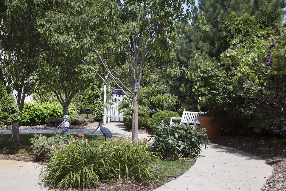 A landscaped outdoor garden path with trees, shrubs, metal bird statues and a white bench near a white picket gate.