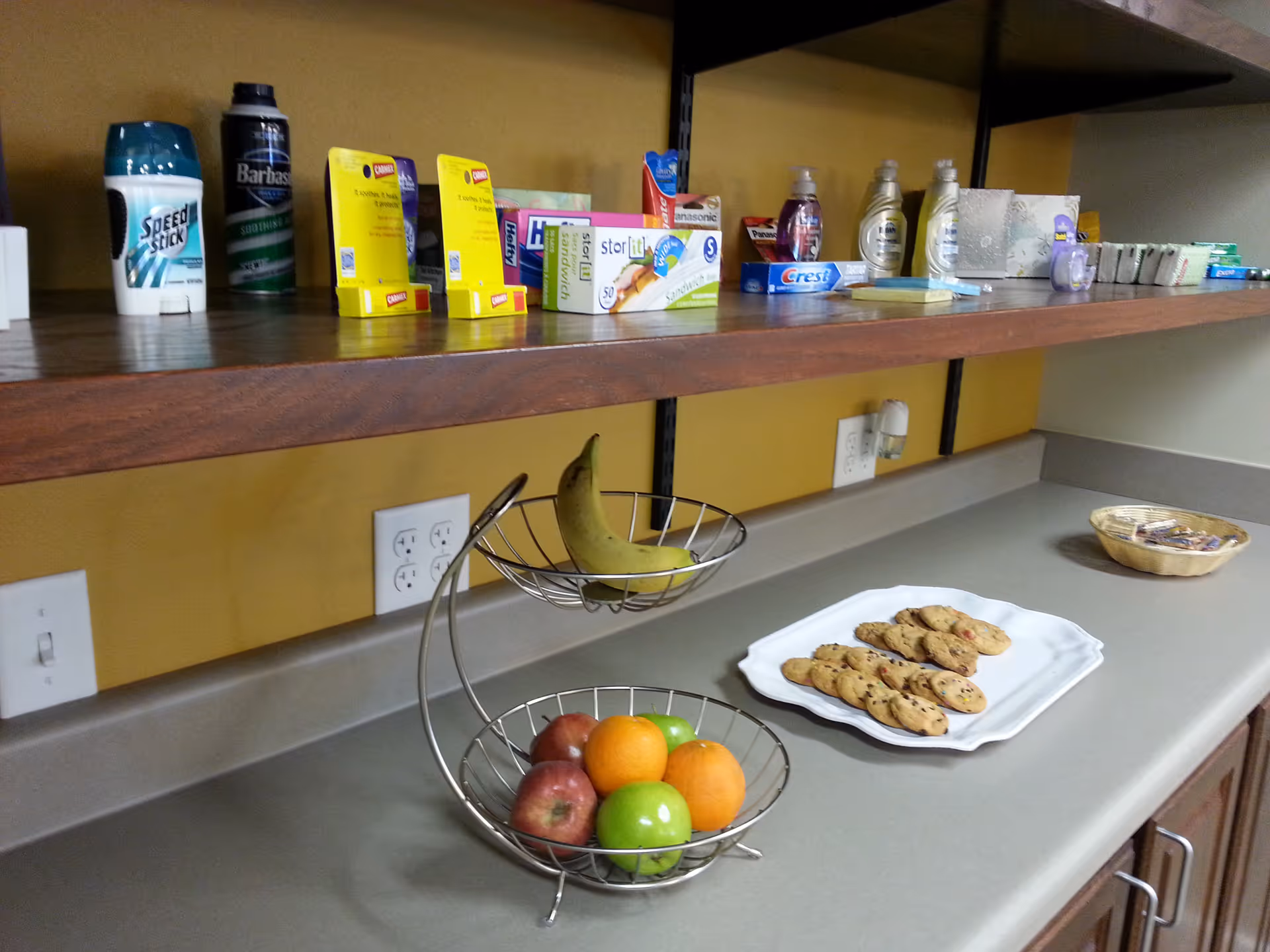 A kitchen counter with a two-tier fruit basket containing bananas, apples, and oranges. On the counter, there is a white plate with chocolate chip cookies and a small basket with individually wrapped snacks. Above the counter, a wooden shelf holds various personal care items including deodorant, shaving cream, toothpaste, and soap. The wall behind is painted yellow and has electrical outlets.