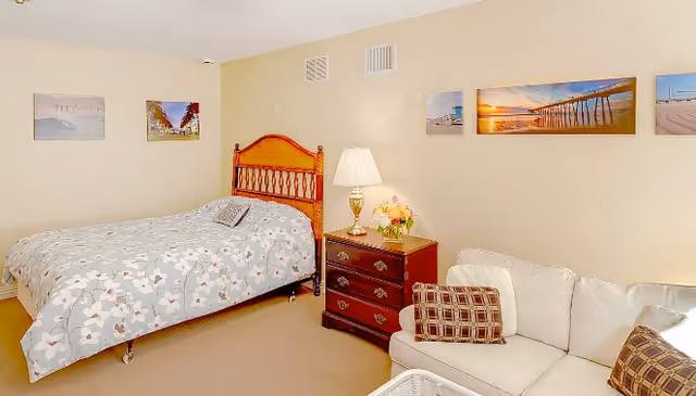 A cozy bedroom with a wooden bed frame and floral bedspread, a wooden nightstand with a lamp and flower arrangement, a white couch with patterned pillows, and several framed pictures of beach scenes on the beige walls.