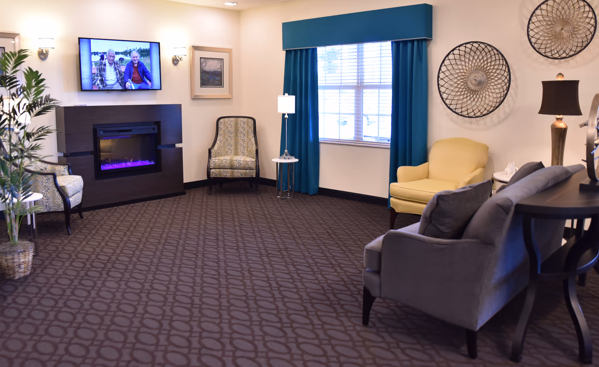 Bright communal lounge with multiple chairs and sofas, a wall-mounted TV above an electric fireplace, teal curtains at a window, and decorative wall art.