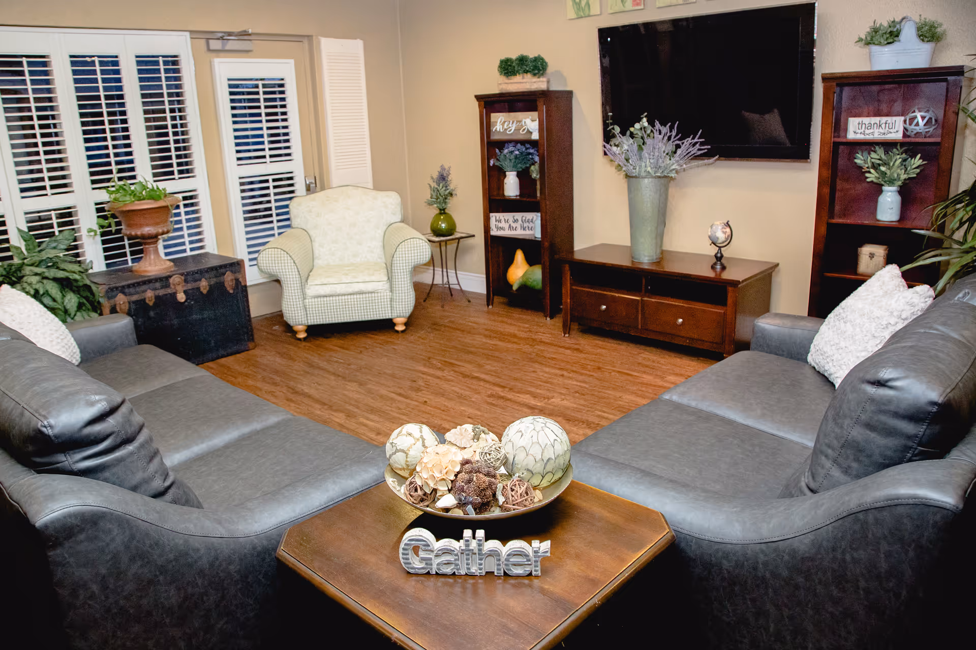 A cozy living room with two dark gray leather sofas facing each other, a wooden coffee table with decorative items including a bowl of natural elements and a 'Gather' sign. There is a light-colored armchair near a set of white French doors with shutters. The room has wooden flooring, two wooden shelving units with plants and decorative signs, a wooden TV stand with a large flat-screen TV mounted on the wall above it, and various plants and decor throughout.
