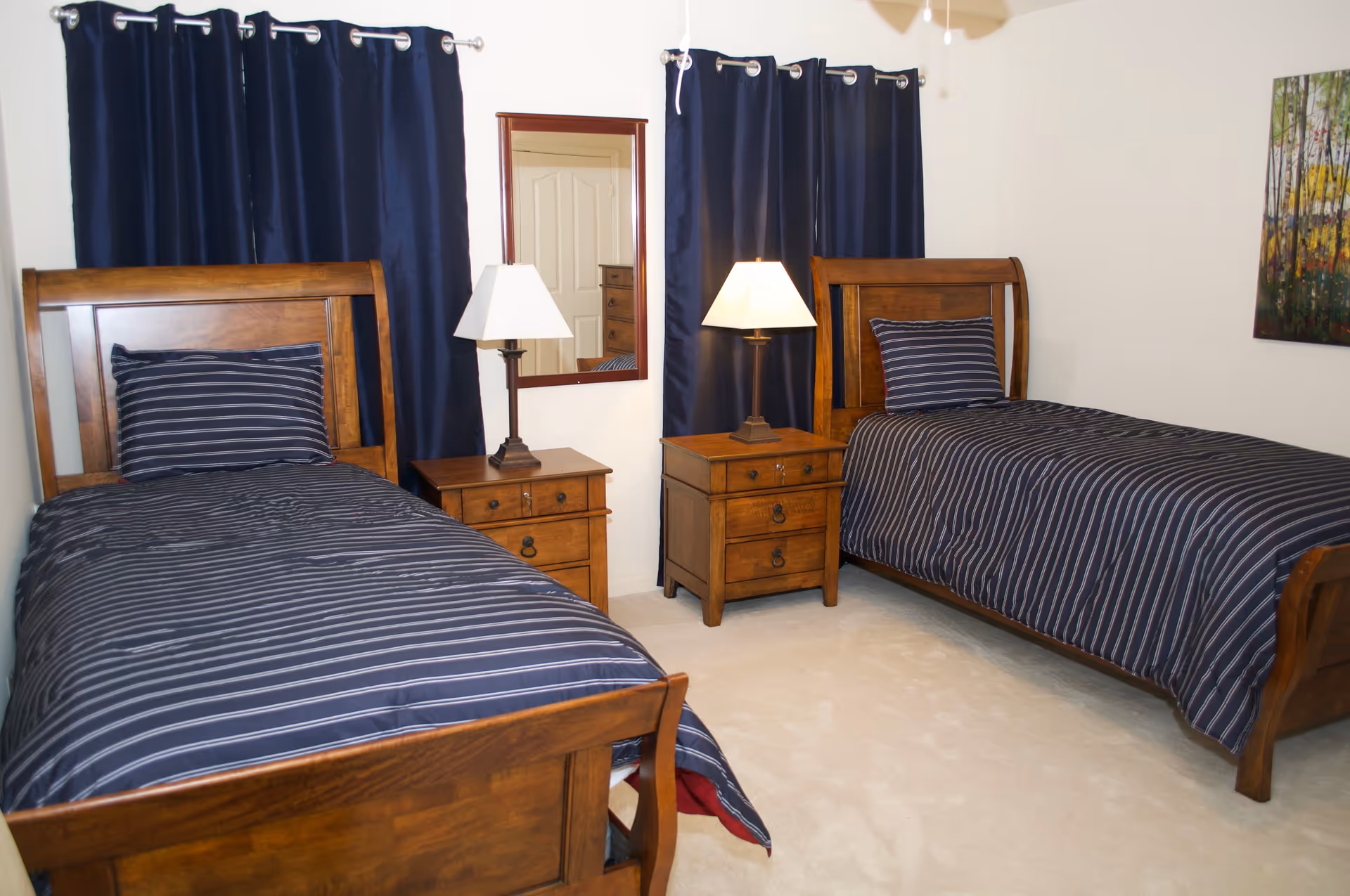 A bedroom with two twin wooden beds, each with navy blue and white striped bedding and matching pillows. Between the beds are two wooden nightstands, each with a lamp. Behind the nightstands are two windows covered with dark blue curtains. A mirror is mounted on the wall between the windows, and a painting of a forest scene hangs on the right wall. The room has light-colored carpet and white walls.