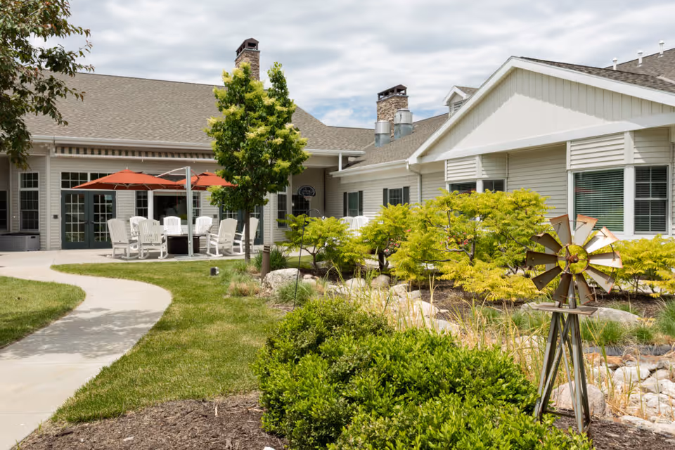Outdoor patio area at CountryHouse Residence featuring a curved concrete walkway, green lawn, shrubs, and a small decorative windmill. White chairs and tables with red umbrellas are arranged on the patio in front of a beige building with multiple windows and chimneys under a partly cloudy sky.