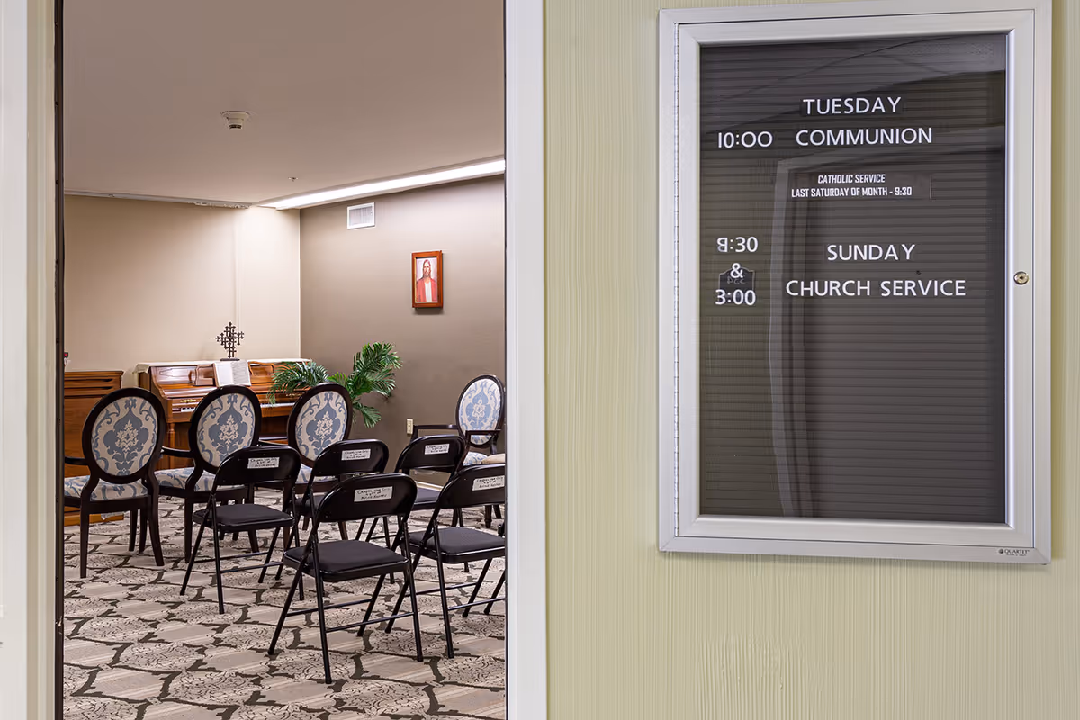 View through a doorway into a small chapel or meeting room with rows of folding and upholstered chairs, a piano, a wall portrait, and a bulletin board showing church service times.