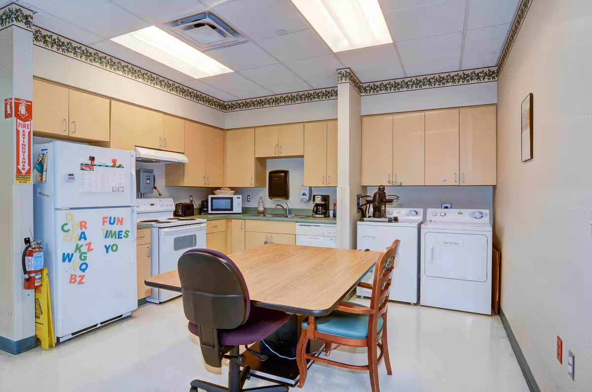 A multipurpose room with a kitchen and laundry area. The kitchen has light wood cabinets, a white refrigerator with colorful alphabet magnets, a white stove, microwave, toaster, coffee maker, and a sink. The laundry area includes a white washing machine and dryer. In the center, there is a wooden table with two chairs, one office chair and one wooden chair with a cushion. The room has a floral border wallpaper near the ceiling and fluorescent lighting.