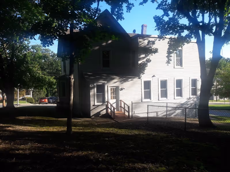 A two-story white residential building partially shaded by trees with a small fenced yard and a wooden ramp leading to a side door. The scene is outdoors with clear skies and sunlight casting shadows from the trees.