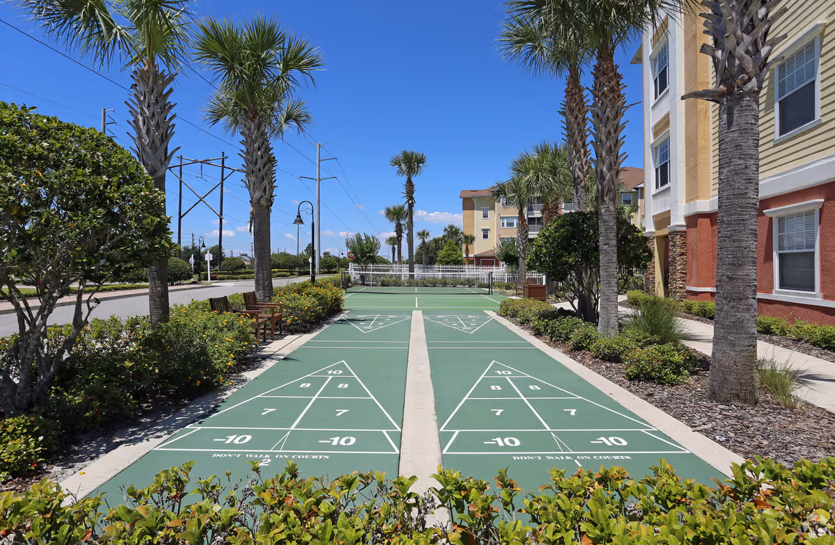 Shuffleboard courts flanked by palm trees and landscaping beside a multi-story residential building under a clear blue sky.
