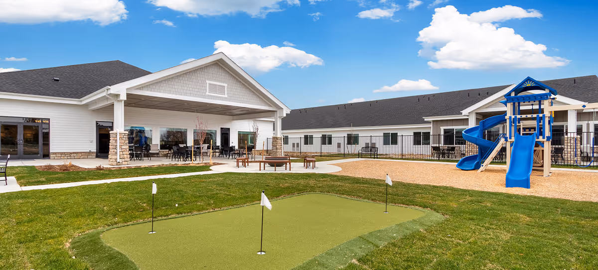 Outdoor area of Greeley Village featuring a small putting green with three flags, a playground with blue slides, a fire pit surrounded by benches, and a covered patio with tables and chairs attached to a white building under a blue sky with clouds.