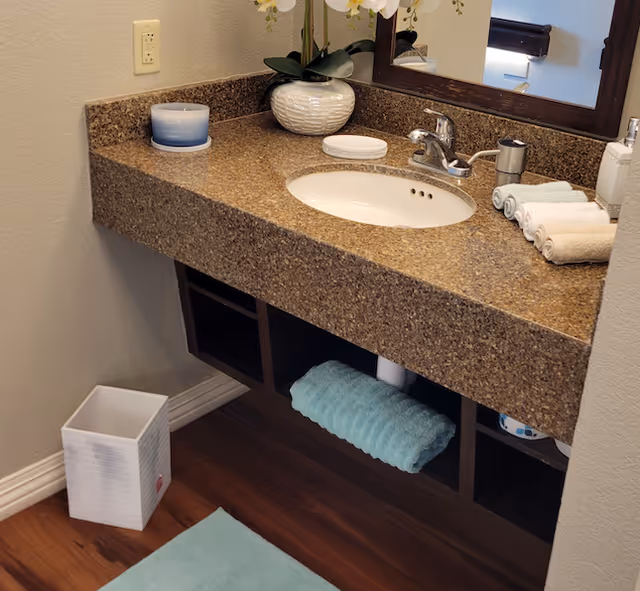 Bathroom vanity with a brown speckled countertop, undermount sink and faucet, mirror, decorative plant, rolled towels, and a trash bin on wood flooring.
