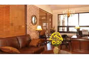 Cozy interior lounge featuring a leather sofa, wooden blinds and brick accent wall, a desk with lamps, and a vase of sunflowers on a table.