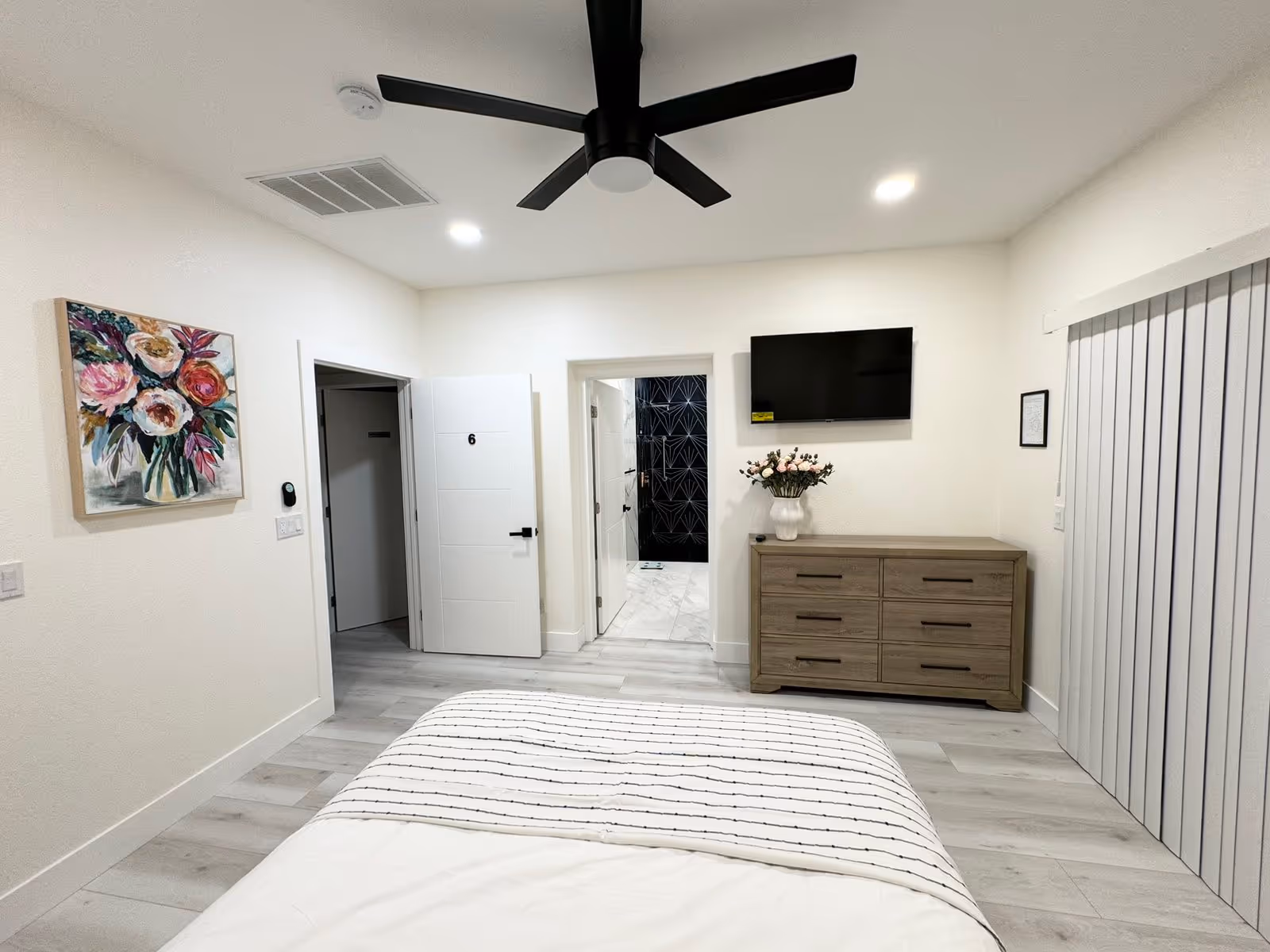 A modern senior care bedroom with light-colored wood flooring, a bed with white and striped bedding, a wooden dresser with a vase of flowers, a wall-mounted TV, a ceiling fan, and two doors leading to other rooms including a bathroom with black and white tile.