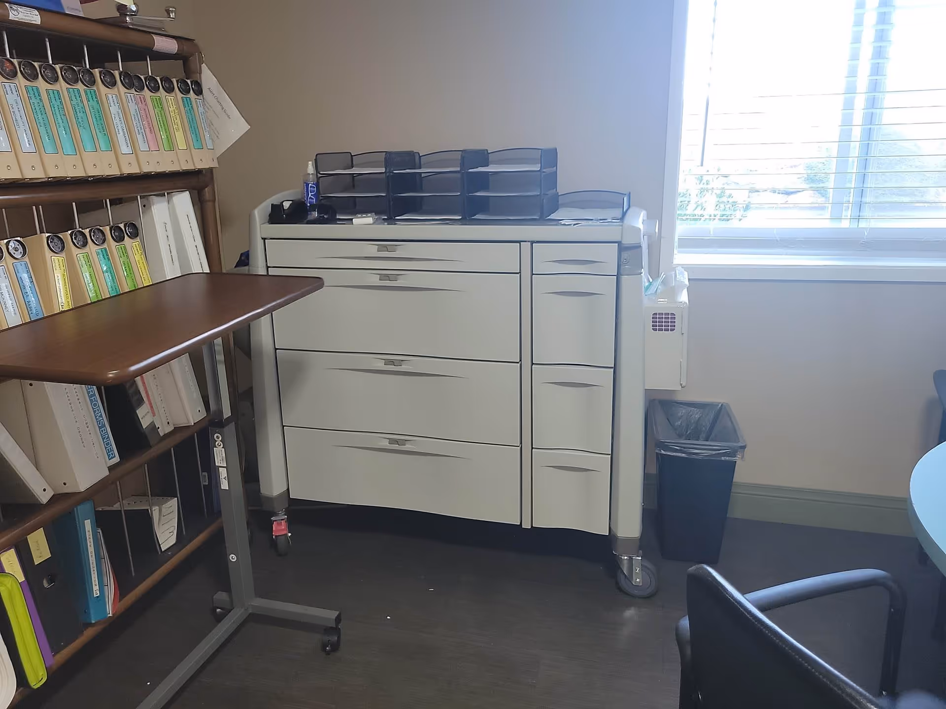 An office corner with a filing cabinet on wheels, a small rolling table, a bookshelf filled with binders, a black trash bin, and a window with blinds letting in daylight.