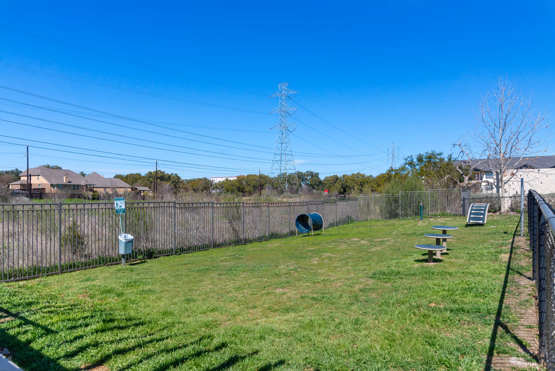 A fenced grassy outdoor area with small play equipment and a tunnel, houses and power lines visible in the background under a blue sky.