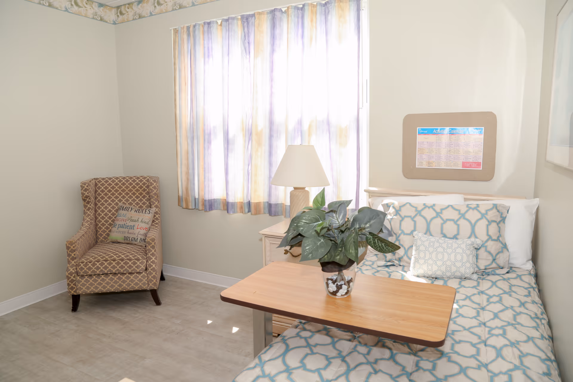 A small, bright senior living bedroom with a single bed, bedside table and lamp, patterned armchair, and a plant on a movable overbed table.