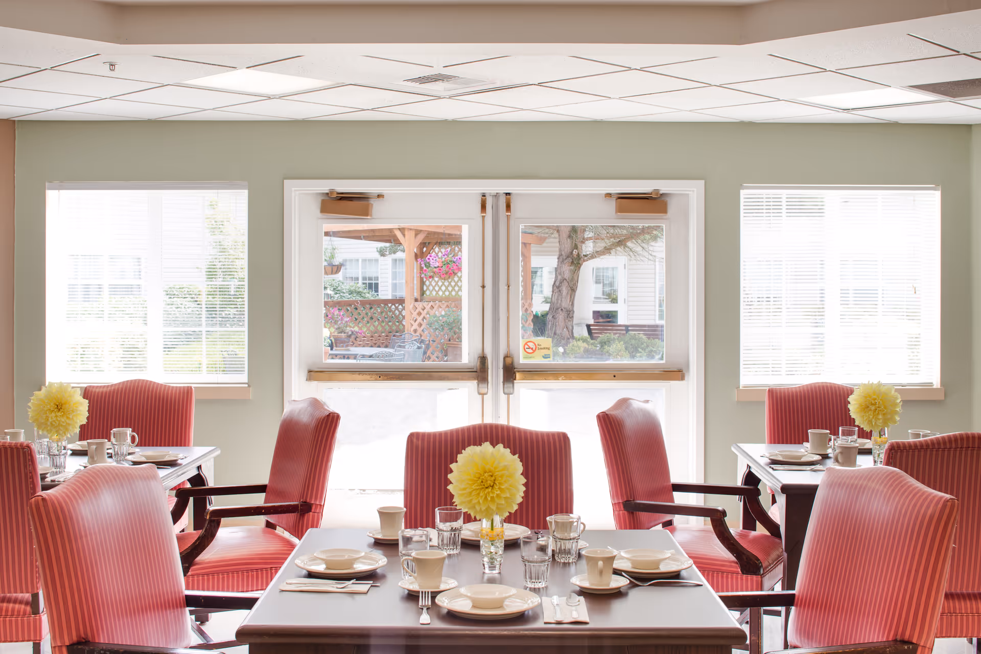 A bright dining room with several tables set for a meal. Each table has red striped chairs and a yellow flower centerpiece. Large windows and glass doors let in natural light and offer a view of an outdoor patio area with hanging flower baskets.