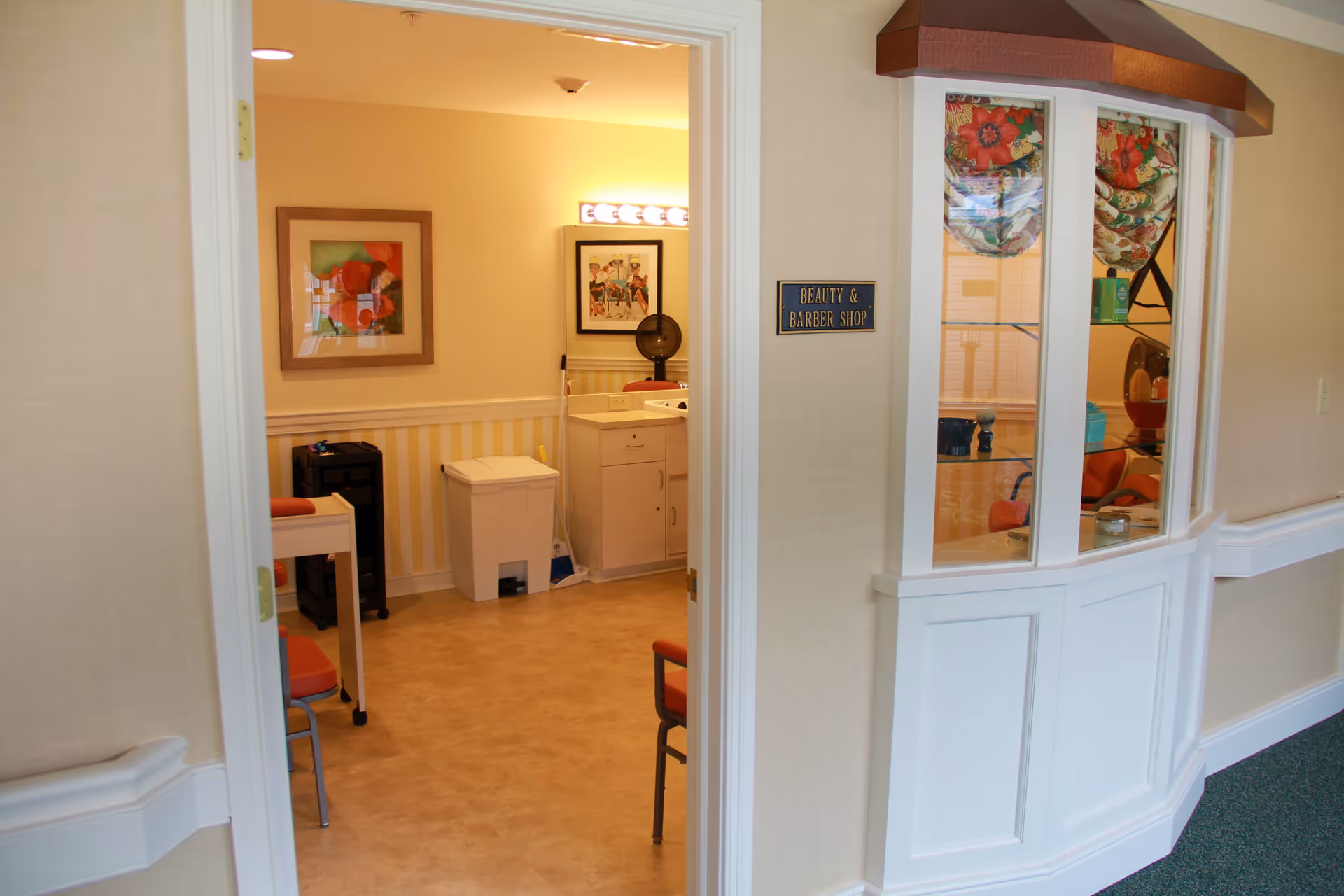 Interior view of a beauty and barber shop in a senior living facility. The room has a counter with a mirror and light bulbs above it, a white trash bin, a small black storage unit, and chairs with orange cushions. The walls are light-colored with framed artwork. A sign outside the room reads 'Beauty & Barber Shop'.