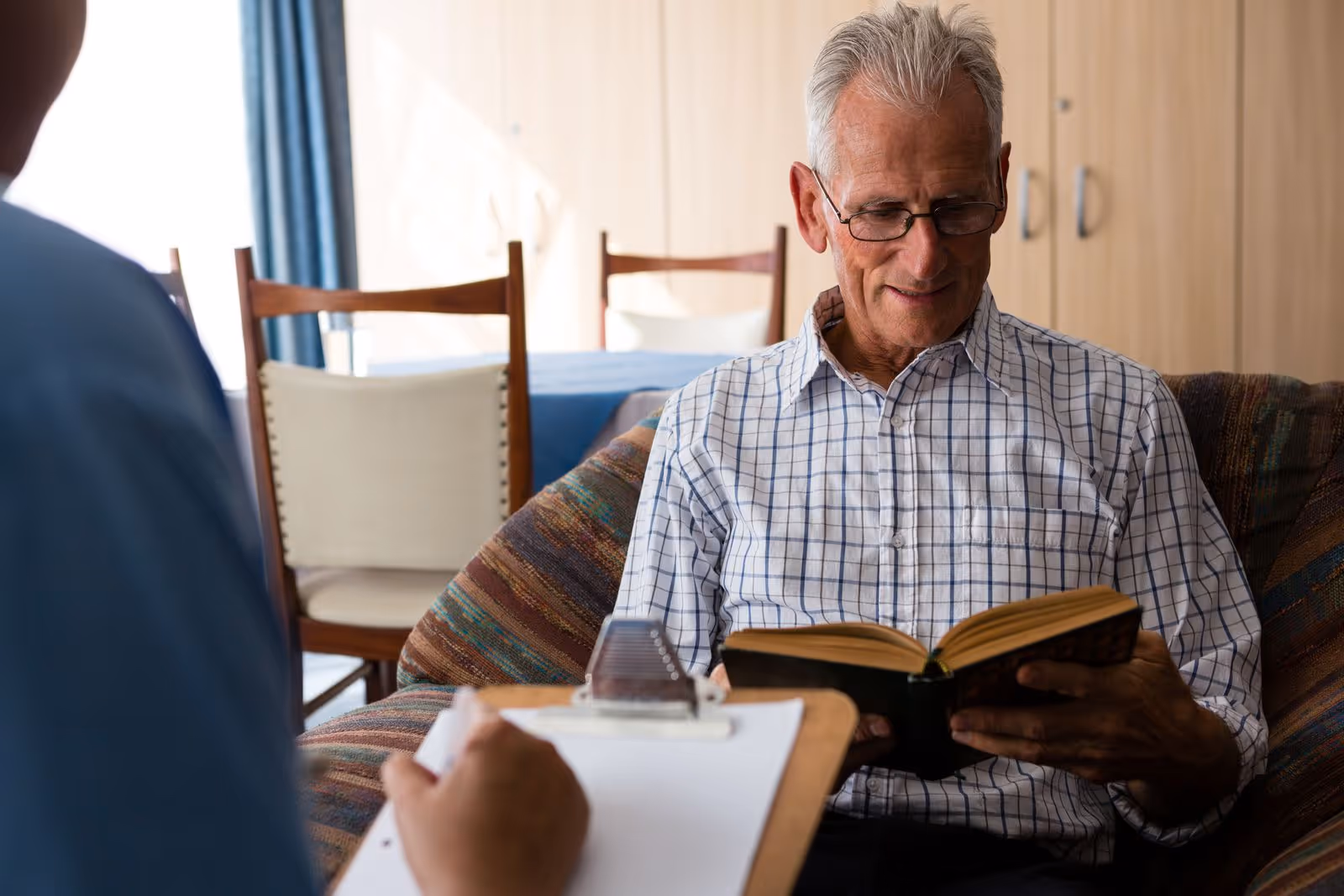 An elderly man wearing glasses and a checkered shirt is sitting on a patterned couch reading a book. In the foreground, a person holding a clipboard is partially visible. Behind them, there is a dining table with chairs and a cabinet in a well-lit room.