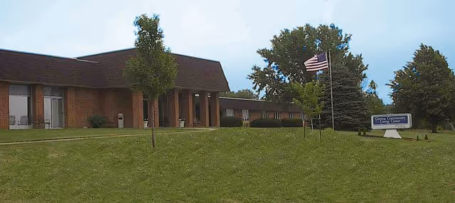 Single-story brick building front with a grassy lawn, flagpoles flying an American flag, trees, and a sign near the entrance.