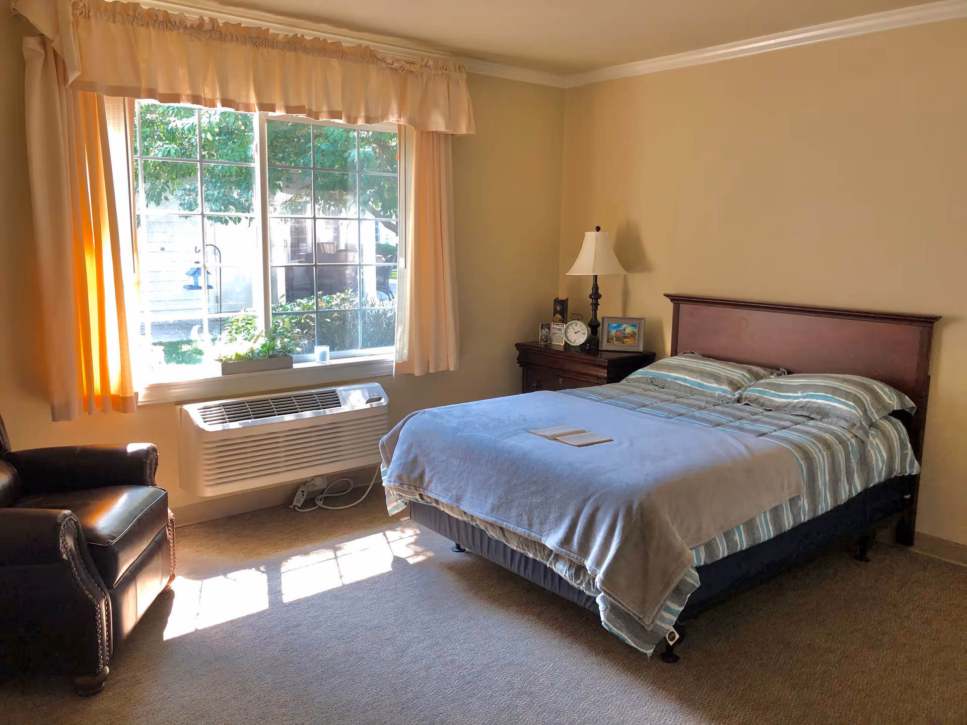 A cozy bedroom with a large window letting in natural light. The room features a bed with striped bedding and a light blue blanket, a wooden nightstand with a lamp, clock, and framed pictures, and a dark leather armchair near the window. Outside the window, greenery and patio furniture are visible.